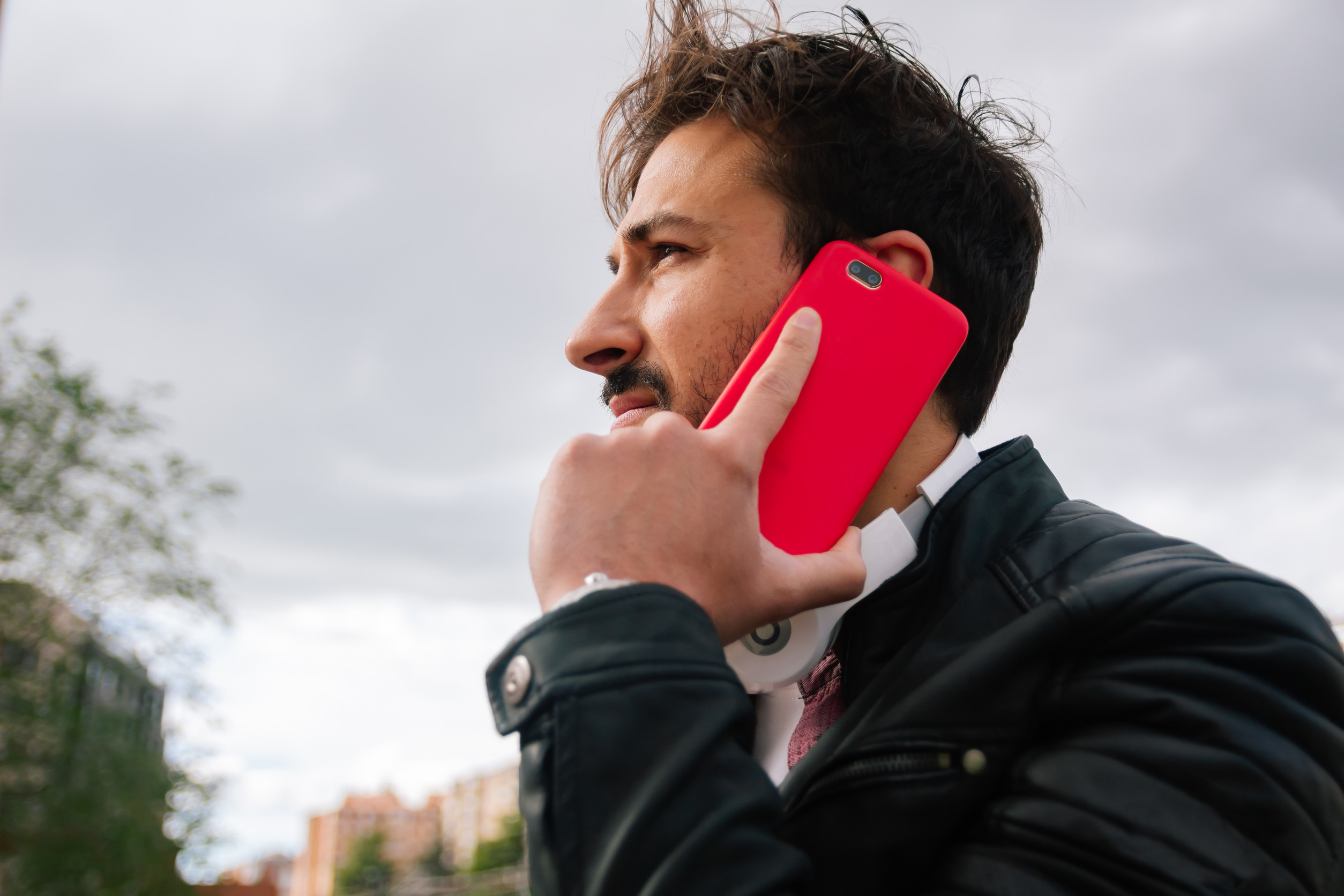 A man outdoors, holding a bright red smartphone to his ear as if he’s on a call. His hair is windblown, he’s wearing a dark jacket, and the cloudy sky and blurred buildings/trees sit in the background.