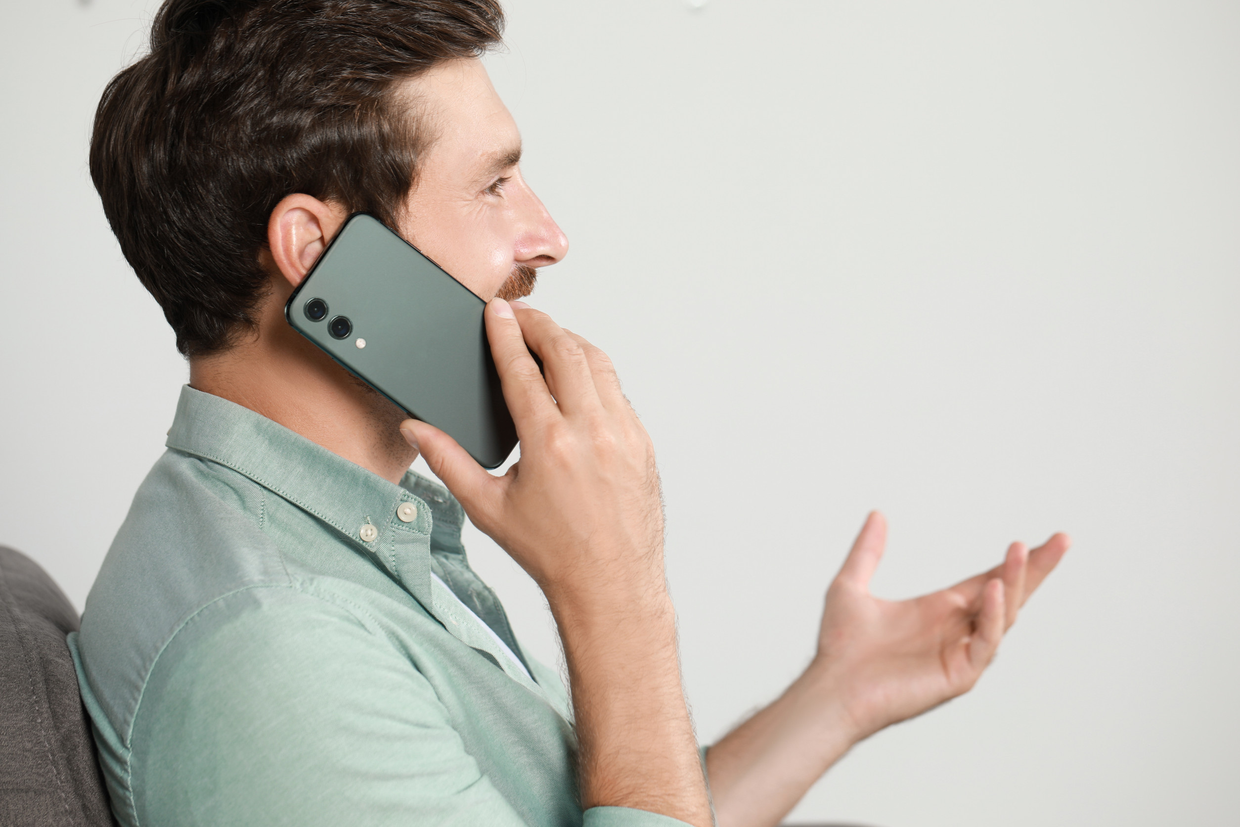 A man in a light green button-down sits indoors, holding a smartphone to his ear while gesturing with his free hand. He’s turned in profile against a plain, bright wall, like he’s mid-conversation. It reads like the moment someone calls because they couldn’t find the price, so the “discovery call” turns into objection handling.