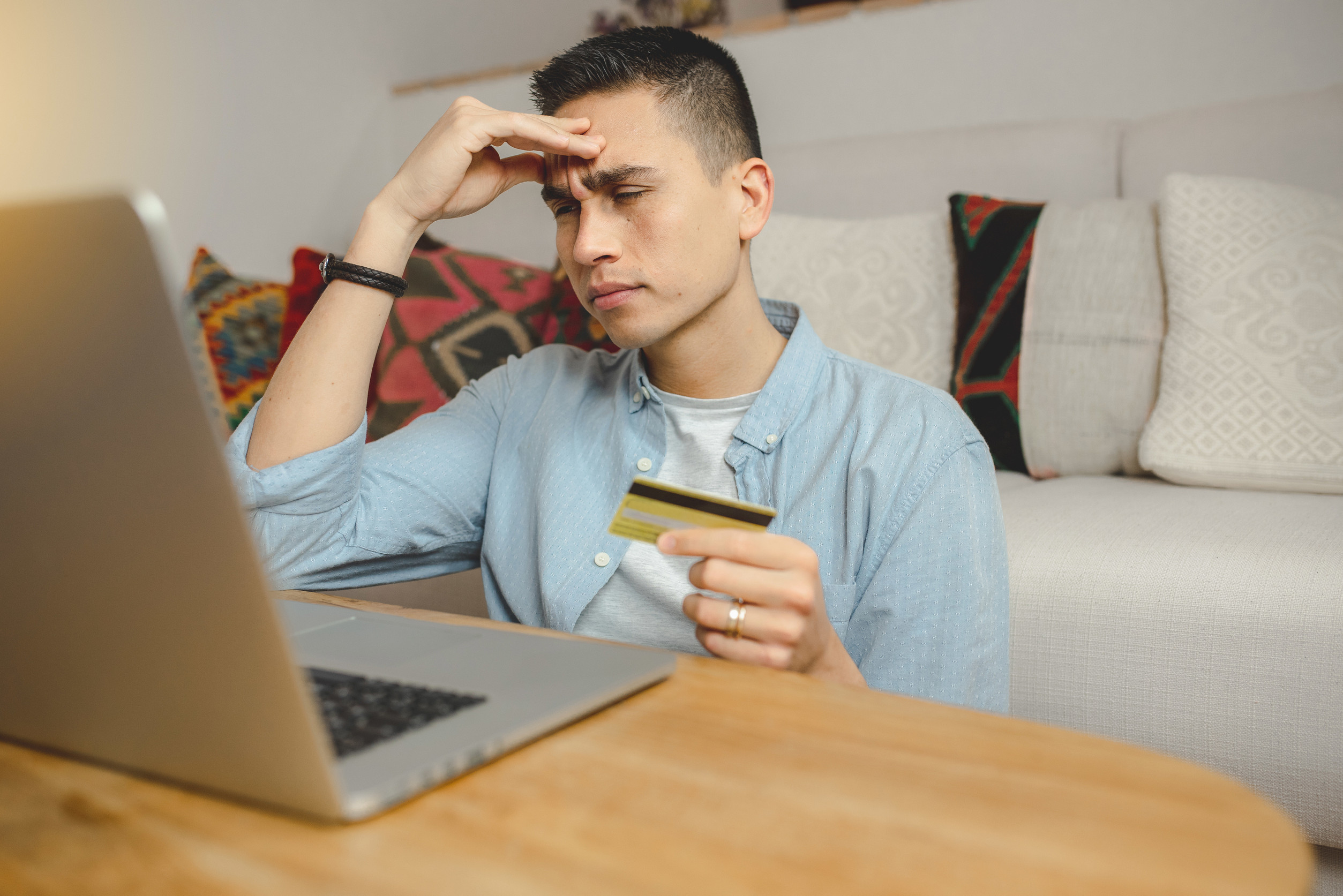 A young man with short dark hair sits at a wooden table in a cozy living room, an open laptop in front of him. He holds a credit card loosely in one hand and presses the other hand against his forehead, his eyes downcast, clearly hesitating before committing to an online coaching offer or digital program.