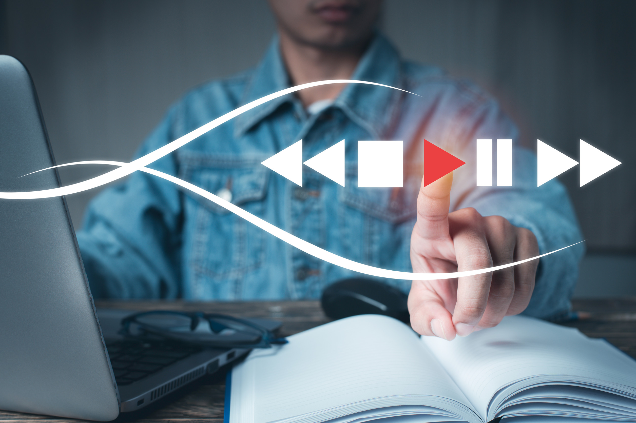 A person at a desk presses a red play button floating over an open notebook and laptop, with rewind/pause/fast-forward icons around it. It looks like someone stopping the “script” and choosing what to say next on purpose.